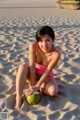 A woman in a pink bikini sitting on a beach with a coconut.